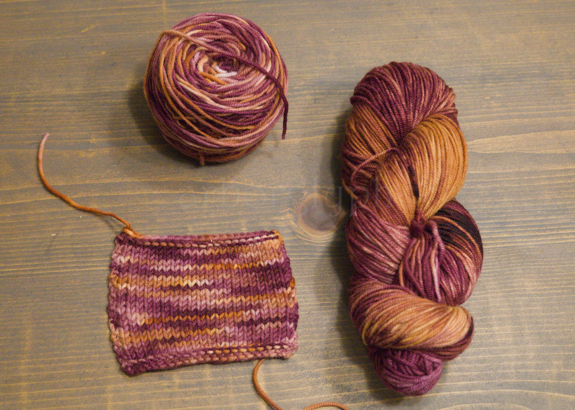 Two skeins of multicolored burgundy and copper yarn and a knitted swatch on a wooden surface.