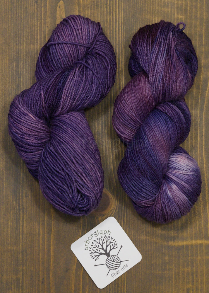 Two skeins of purple yarn on a wooden surface with a brand label.