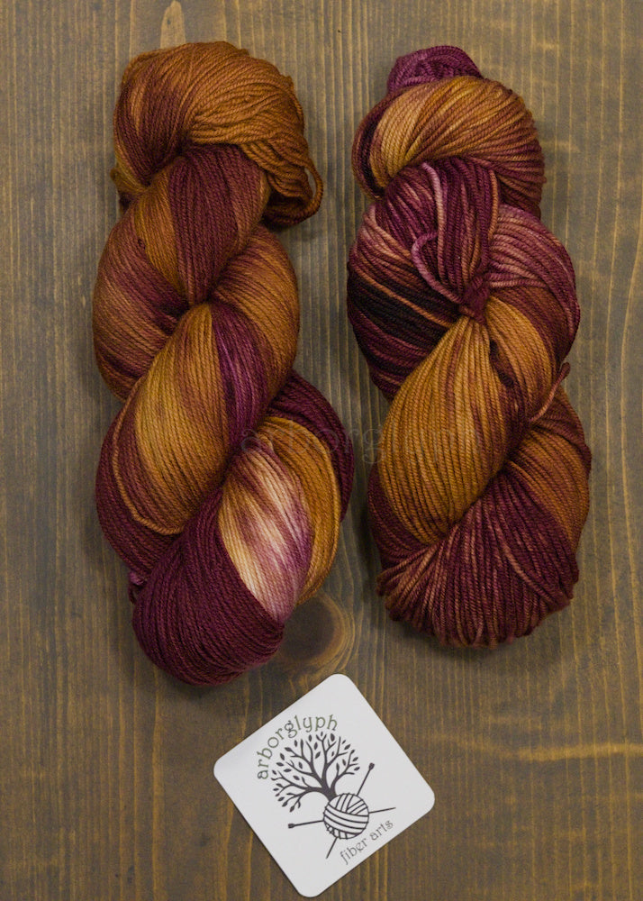 Two skeins of multicolored yarn, burgundy and copper, on a wooden surface with a brand label.