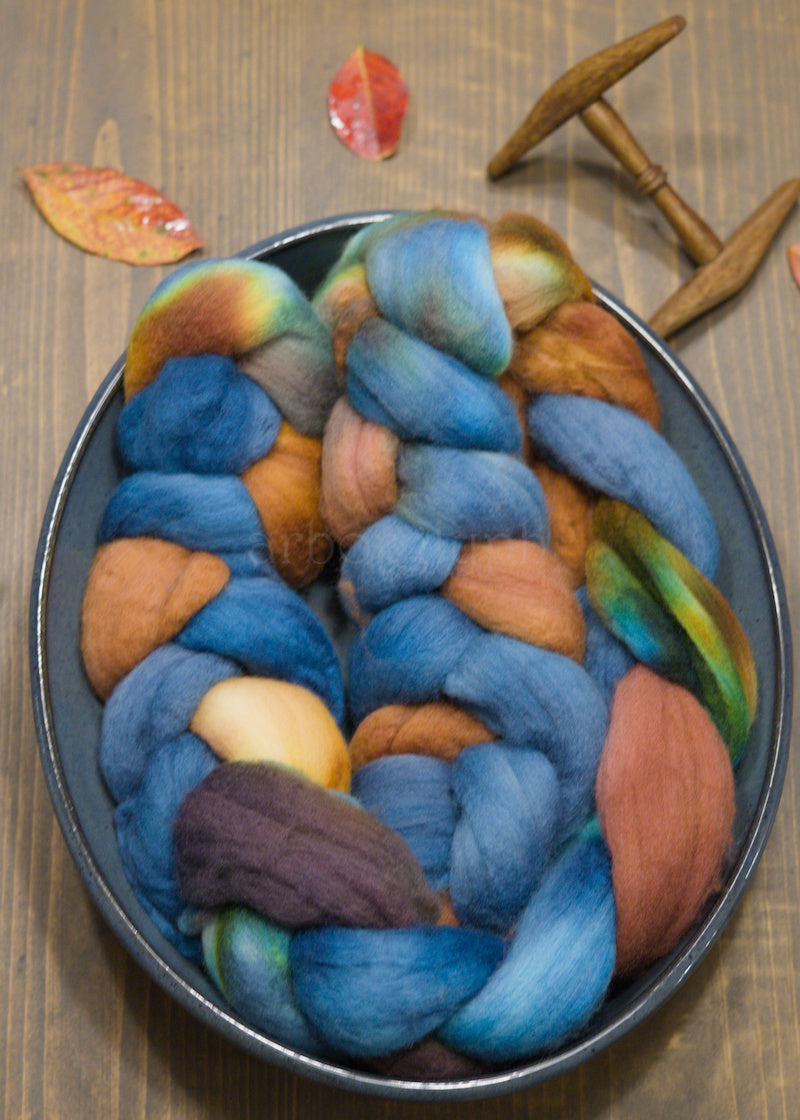 Blue and copper combed top in a bowl on a wooden surface with leaves and a spindle.