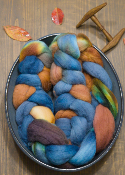 Blue and copper combed top in a bowl on a wooden surface with leaves and a spindle.