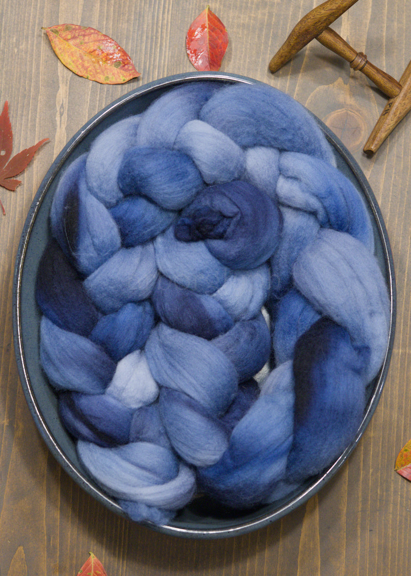 Bowl filled with blue combed wool top on a wooden surface with leaves and a spindle.