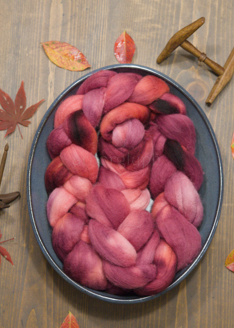 Braided Merino combed top of pink and red yarn in a bowl on a wooden surface with autumn leaves.