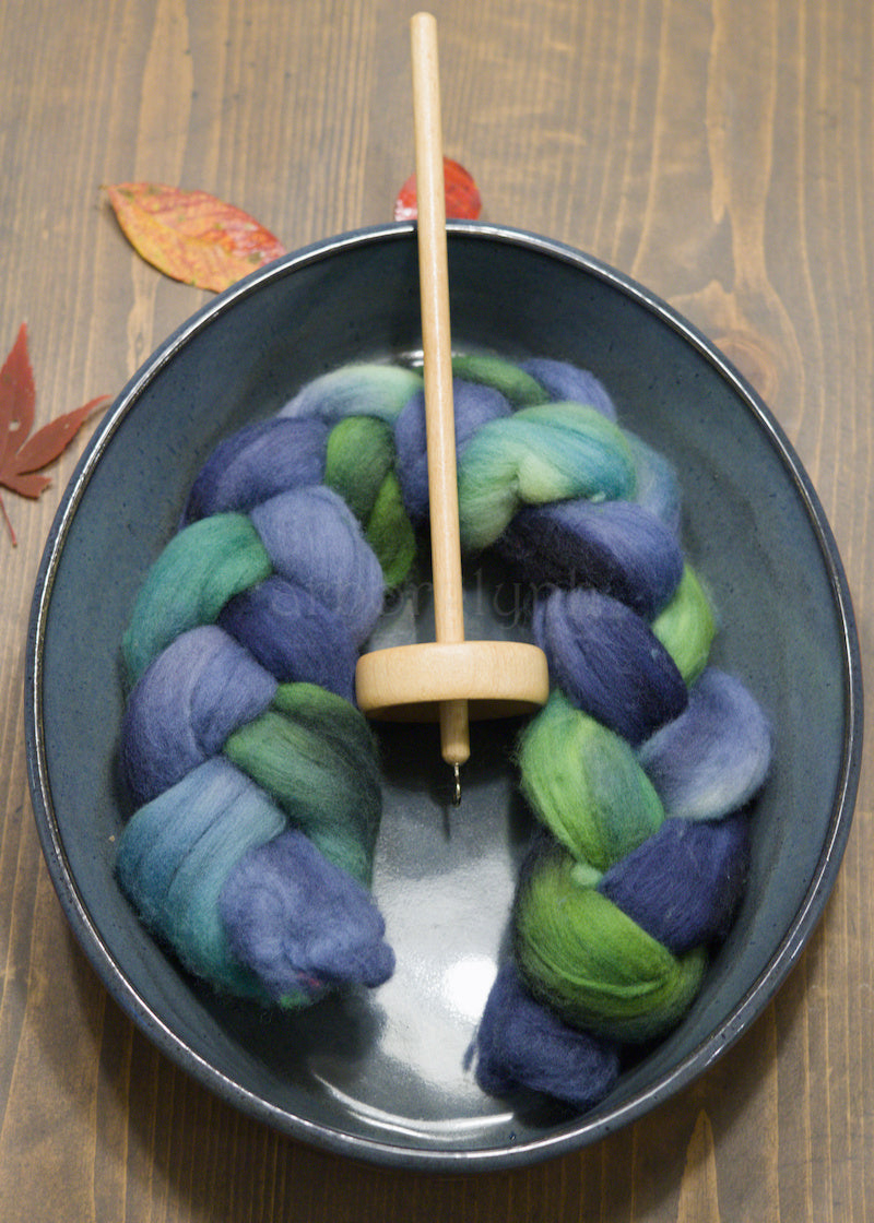 Drop spindle with blue and green combed wool top in a bowl on a wooden surface