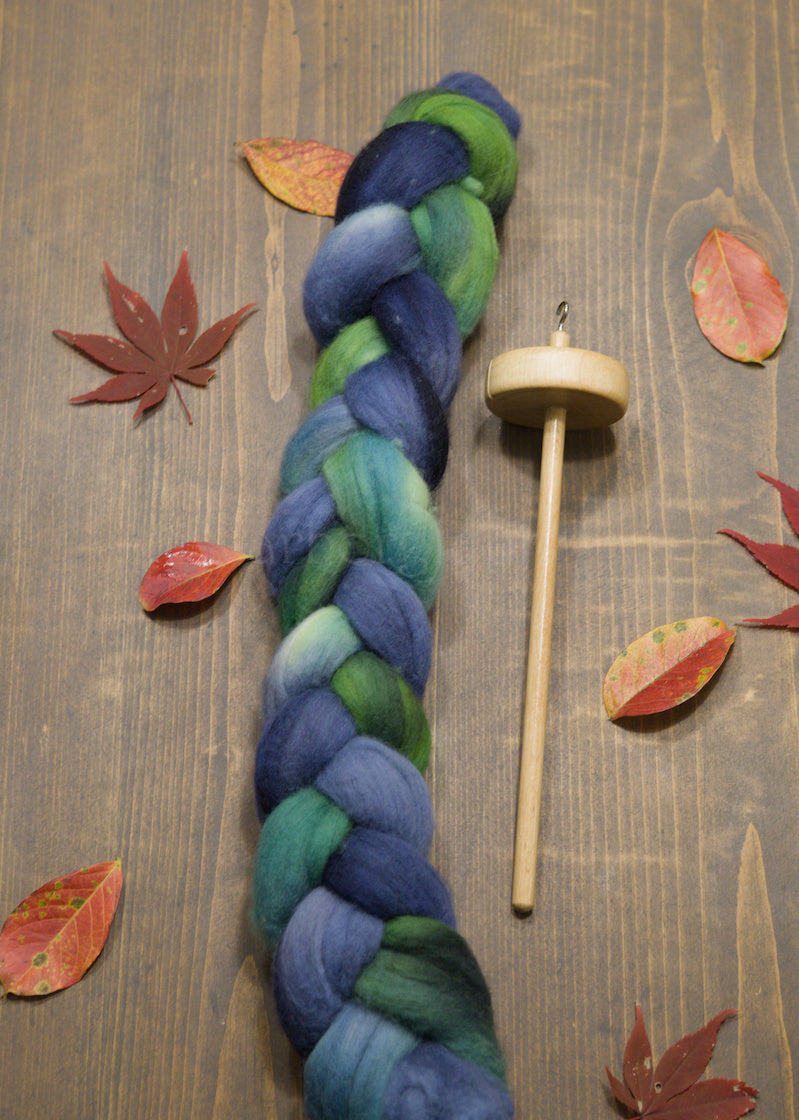 Braided blue and green combed wool top with a wooden spindle on a wooden surface with fall leaves.