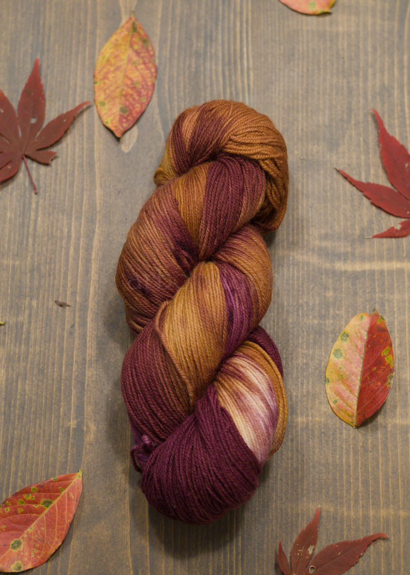 Multicolored, burgundy and copper, skein of yarn on a wooden surface with autumn leaves.